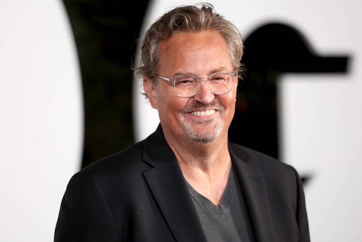 Matthew Perry smiles while wearing a gray tshirt and black jacket